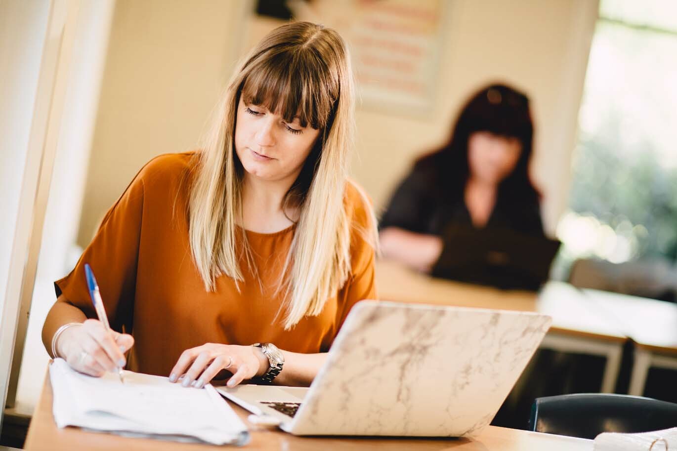 Student writing in notebook whilst studying from laptop